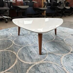 A used mid-century modern triangular coffee table featuring a high-gloss white lacquered top and three tapered solid walnut legs, displayed on a blue geometric area rug in a modern office lounge setting.